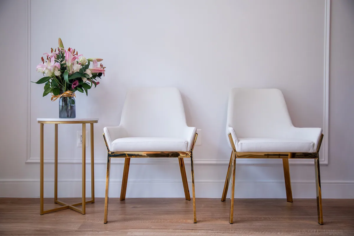 Pair of white chairs with warm metal frames beside a small flower table.